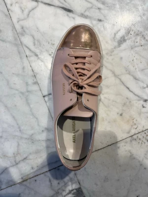 Rosa sneakers Axel Arigato - Snygga sneakers från Axel Arigato i ljusrosa skinn med metallic guldigt tåparti. Klassisk låg modell med vita sulor och snörning framtill. Perfekta för dig som vill ha en clean men ändå unik look.
