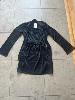 Svart omlottklänning från BikBok - Stilren svart omlottklänning från BikBok i blankt material. Klänningen har lång ärm, markerad midja och klassisk omlottdesign som ger en snygg siluett. Perfekt för dig som gillar enkel men elegant stil.