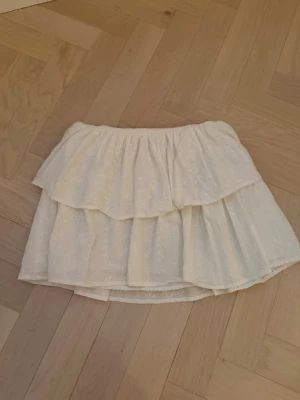 Vit volangkjol med broderade detaljer - Söt vit kortkjol med dubbla volanger och broderade små blommor. Kjolen har resår i midjan för skön passform och är gjord i ett lätt bomullstyg, perfekt för varma dagar. Passar dig som gillar romantisk och feminin stil.