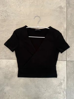 Svart omlottopp från even&odd  - Svart omlottopp med djup v-ringning, korta ärmar och croppad passform, från Even&odd💕 Köptes från Zalandos hemsida tillsammans med en likadan vit (Nypris 2st 220kr). Säljer för att den aldrig kommit till användning, så den är helt som ny!🩷 (Står ej för frakt och pris kan diskuteras)