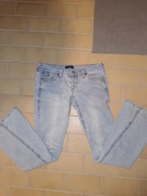 TRUE religion ljusblå low waist / lågmidjade flared bootcut  - Säljer ett par ljusblå jeans från True Religion med klassiska broderade bakfickor och silverfärgade knappar. Jeansen har rak passform och femficksdesign. Perfekta för dig som gillar streetstyle och vill sticka ut med snygga detaljer.