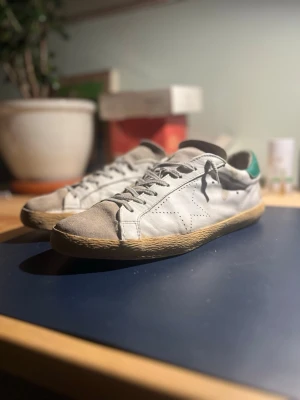 Vita GGDB SSTAR sneakers  - Säljer ett par vita sneakers från GGDB SSTAR med klassisk låg profil, snörning och stjärndetalj på sidan. Skorna har grå/gröna detaljer och en beige sula. Tillverkade i skinn och har en stilren, avslappnad look. Jävligt ovanliga pga att dem var väldigt limited och såldes för länge sedan, går knappt att hitta