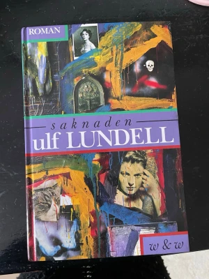 Saknaden av Ulf Lundell - En färgstark roman av Ulf Lundell med ett konstnärligt omslag i lila, gult, blått och rött. Boken är inbunden och har collage-liknande bilder på framsidan. Perfekt för dig som gillar svensk samtidslitteratur och starka berättelser.