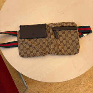 Gucci beige magväska med GG-mönster - Snygg beige magväska från Gucci med klassiskt GG-mönster och bruna detaljer. Väskan har två fack framtill, ett med dragkedja och ett med lock, samt ett extra fack på baksidan. Justerbart band i grönt och rött med metallspänne.