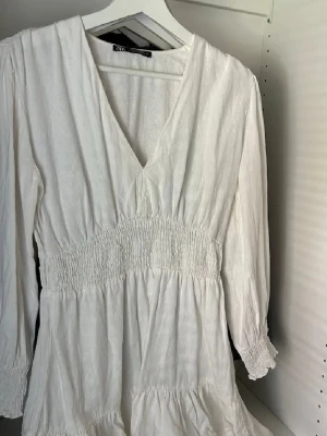 Kort Vit klänning  - Superfin vit kortklänning från ZARA med v-ringning och smock i midjan. Klänningen har långa ärmar med smock vid ärmslut och volang nertill. Perfekt för sommarens alla tillfällen och som studentklänning.Skriv gärna vid frågor!💕