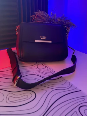 Svart axelväska från Guess - Snygg svart axelväska från Guess med guldfärgade detaljer och bred axelrem. Väskan har en stilren design med logotyp framtill och dragkedja upptill. Perfekt för dig som vill ha en trendig och praktisk väska till vardagen.