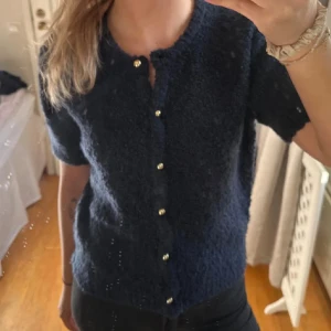 Mörkblå kortärmad tröja Gina Tricot - Säljer en mörkblå, kortärmad kofta från Gina Tricot med guldknappar framtill. Koftan har en mjuk, fluffig struktur och rund halsringning. Knappt använd! 