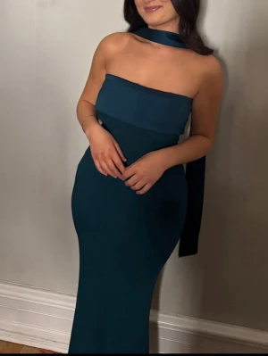 Smaragdgrön axelbandslös långklänning - Elegant smaragdgrön långklänning med axelbandslös design och en bred scarf-detalj runt halsen. Klänningen har en figurnära passform och är perfekt för speciella tillfällen. Materialet ser ut att vara mjukt och följsamt, vilket ger en snygg siluett.