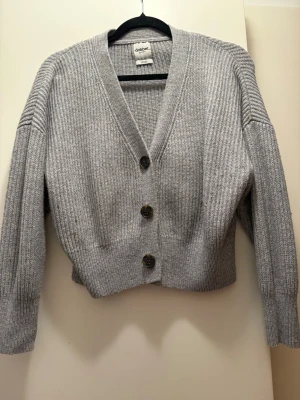 Grå ribbstickad kofta från Dobber - Snygg grå ribbstickad kofta från Dobber med tre stora knappar framtill. Koftan har v-ringning, långa ärmar och en croppad passform. Perfekt att slänga över en t-shirt eller linne för en chill look.