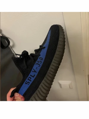 Yeezy Boost 350 V2 svart/blå - Säljer ett par Yeezy Boost 350 V2 med svart Primeknit-överdel och blå sidostripes med texten SPLY-350. Skorna har en chunky, räfflad sula i mörkgrått och klassisk snörning. Perfekta för dig som gillar streetwear och vill sticka ut.