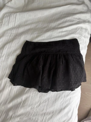 Svart kortkjol med volang - Säljer en svart kortkjol med volang och diskreta små prickar. Kjolen har en bred midjeresår och ett lätt, luftigt tyg som ger en snygg siluett. Perfekt att matcha med en enkel topp eller stickad tröja.