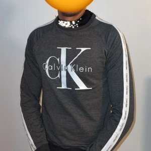 Mörkgrå Calvin Klein sweatshirt - Snygg mörkgrå sweatshirt från Calvin Klein med stort CK-tryck på bröstet och vita ränder med logga längs ärmarna. Tröjan har rund halsringning och en avslappnad passform. Perfekt för dig som gillar streetwear och vill ha något stilrent.