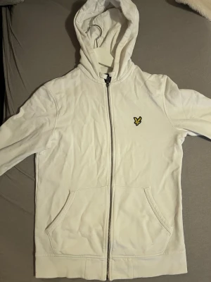 Vit hoodie från Lyle & Scott - Säljer en vit hoodie från Lyle & Scott med dragkedja framtill och klassisk gul logga på bröstet. Hoodien har fickor på magen, ribbade muddar och en skön huva. Perfekt för en avslappnad och stilren look.