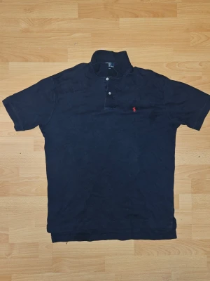 Marinblå piké från Ralph Lauren - Klassisk marinblå pikétröja från Ralph Lauren med korta ärmar och knappar framtill. Ikoniska röda broderade loggan på bröstet. Tillverkad i mjuk bomull för en bekväm känsla och stilren look.