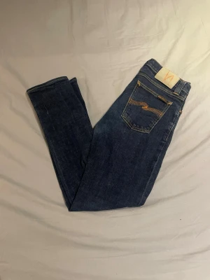 Nudie Jeans blå jeansbyxor W28 L32 - Säljer ett par klassiska blå jeans från Nudie Jeans med snygga kontrastsömmar och ikonisk broderad bakficka. Modellen har normal passform och raka ben. Tillverkade i slitstarkt bomullsmaterial, perfekta för en avslappnad och stilren look.