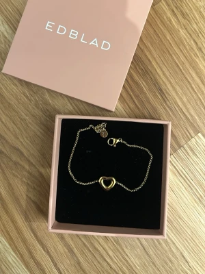 Guldigt armband med hjärta från Edblad - Snyggt och stilrent armband från Edblad i guldfärgat metall med ett öppet hjärta i mitten. Tunt kedjearmband med karbinlås och liten Edblad-tag vid låset. Perfekt accessoar för att lyfta din outfit.