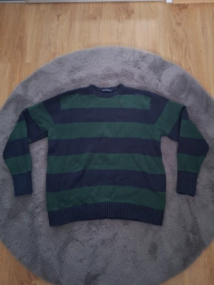 Randig stickad tröja i grönt och marinblått - Säljer en stickad tröja med breda horisontella ränder i mörkgrönt och marinblått. Tröjan har rund halsringning, långa ärmar och ribbstickade muddar vid ärmslut och nederkant. Perfekt för dig som gillar klassisk och avslappnad stil.