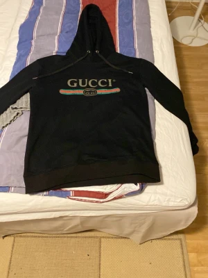 Svart hoodie från Gucci med logga - Säljer en svart hoodie från Gucci med stor logga och färgglad rand på bröstet. Klassisk huva med dragsko och ribbade muddar. Hoodien har en avslappnad passform och är tillverkad i mjukt material, perfekt för en trendig och bekväm stil.