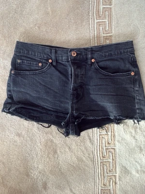Svarta jeansshorts med rå kant - Säljer ett par svarta jeansshorts med rå, fransig kant och klassisk femficksdesign. Shortsen har knapp och dragkedja framtill samt bälteshällor. Perfekta för sommaren och ger en avslappnad look.
