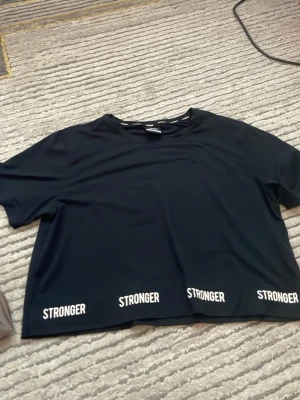 Svart croppad t-shirt från Stronger - Svart croppad t-shirt från Stronger med vit logga tryckt flera gånger längs nederkanten. T-shirten har korta ärmar och är gjord i ett mjukt, stretchigt material som passar perfekt till en aktiv livsstil, luftig också!