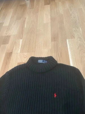 Svart stickad tröja Polo Ralph Lauren - Klassisk svart stickad tröja från Polo Ralph Lauren med ribbad struktur och hög krage. Tröjan har lång ärm och den ikoniska röda logotypen broderad på bröstet. Perfekt för dig som gillar stilrena och tidlösa plagg.