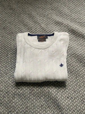 Vit kabelstickad tröja Morris  - Vit kabelstickad tröja från Gant med rund halsringning och diskret blå logga broderad på bröstet. Tröjan har långa ärmar och ribbade muddar vid ärmslut och nederkant. Perfekt för en klassisk och stilren look.