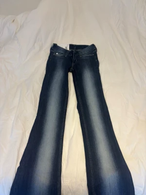 Blå bootcut jeans från weekday - Säljer ett par blå bootcut jeans från weekday med snygg slitning längs benen och klassisk femficksmodell. Jeansen har en vit patch med logga bak och är tillverkade i denim med lätt stretch. Perfekta för en avslappnad men trendig look.