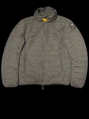 Mörkgrön dunjacka från Parajumpers - Snygg grå dunjacka från Parajumpers med hög krage och gul detalj vid halsen. Jackan har två sidofickor med dragkedja och en patch med logga på ena ärmen. Perfekt för kalla vinterdagar och riktigt stilren look.
