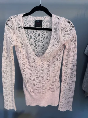 Ljusrosa virkad stickad tröja - Söt ljusrosa stickad tröja med virkat mönster och djup urringning. Tröjan har långa ärmar och bred ribbad mudd nedtill. Materialet är tunt och genombrutet, perfekt för lager på lager. Märket syns på etiketten men är otydligt.