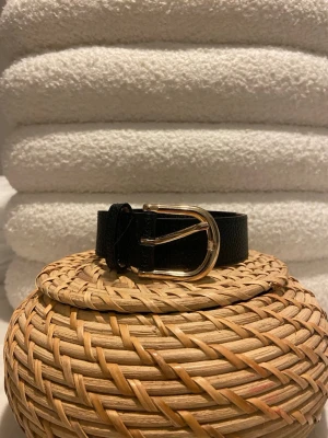 Svart bälte med guldfärgat spänne - Stilrent svart bälte i skinn med guldfärgat, rundat spänne. Perfekt accessoar för att lyfta din outfit och funkar till både jeans och klänning. Bältet har en klassisk design och är lätt att matcha med olika stilar.