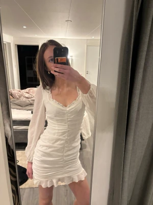 Vit kort omlottklänning med volang - Supersöt vit kort klänning med långa transparenta ärmar, v-ringning och volangdetaljer vid urringning och nederkant. Inte använt denna på väldigt länge därav säljer jag den! Sitter som xs och s! Vet inte exakt var den är ifrån men står ”kiss the sky” i den:)