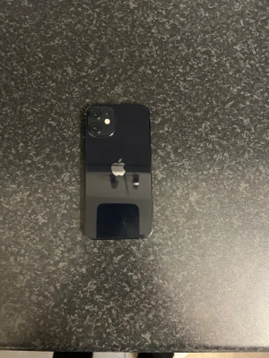 Svart iPhone 12 med dubbla kameror - Snygg svart iPhone 12mini  med stilren design och rundade kanter. Baksidan är i glas och har två kameror samt Apple-loggan i mitten. Telefonen har en platt framsida med stor skärm och tunna ramar. Perfekt för dig som gillar modern teknik och enkel design.