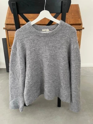 Grå stickad tröja från Gina Tricot - Mysig grå stickad tröja från Gina Tricot med rund halsringning och ribbade muddar vid ärmslut och nederkant. Tröjan har en avslappnad passform och är tillverkad i en mjuk ullblandning som passar perfekt för kyliga dagar.