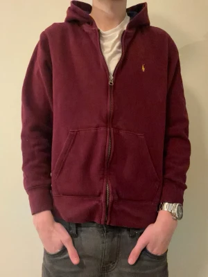 Ralph lauren hoodie - strl 14-16 - Ralph Lauren zip up hoodie 🧥✨ Vinröd färg med gul logga ❤️💛 Storlek 14–16 år, passar modellen som har XS men är lite mindre i passformen 👀 Finns ett litet hål på armen, inget stort och inget som syns mycket 🪡 Snygg och clean hoodie som funkar till många outfits 😎🔥 Hör av er om ni har frågor eller vill ha fler bilder 💬 