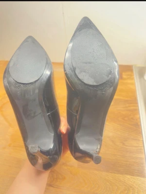 Svarta klassiska pumps i storlek 38 - Säljer ett par svarta pumps med spetsig tå och hög klack. Skorna har en glansig finish och är tillverkade i syntetmaterial. Perfekta för dig som vill ha en stilren och tidlös look.