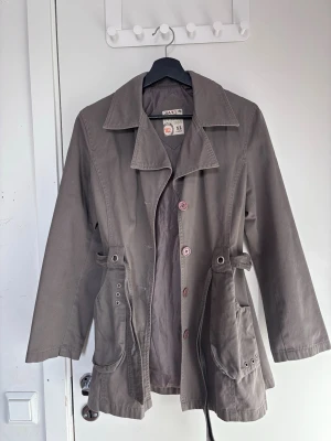 Brun trenchcoat från Shanlin - Den är i storlek xl men passar som M Stilren brun trenchcoat från Shanlin med klassisk krage, knäppning framtill och stora fickor med detaljerade öljetter. Jackan har använts ett par gånger, den har ett justerbart midjeband och är tillverkad i ett slitstarkt bomullsliknande material. Perfekt för dig som vill ha en enkel men snygg look.