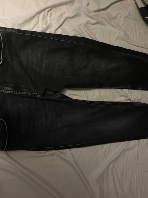 Svarta jeans från Nudie Jeans - Säljer ett par svarta jeans från Nudie Jeans med klassisk femficksdesign och smal passform. Jeansen har snygga kontrastsömmar och en brun läderpatch bak i midjan. Perfekta för dig som gillar stilrena och tidlösa jeans.