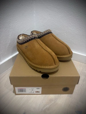  UGG tofflor, tasman - Mysiga bruna tofflor från UGG i mocka. Dekorativt mönster runt öppningen och robust sula som passar perfekt för hemmabruk. Klassisk slip-on modell som är både snygg och bekväm. Tasman modell