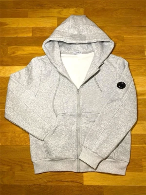 Ljusgrå zip hoodie från C.P. Company - Ljusgrå hoodie med dragkedja från C.P. Company. Klassisk huva med dragsko, två fickor framtill och ribbade muddar. Ikonisk svart logodetalj på ärmen. Tillverkad i mjuk bomullsblandning för skön känsla.