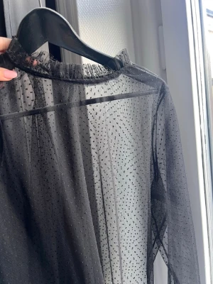 Svart mesh topp från NA-KD - Säljer en  svart mesh topp från NA-KD med små prickar och volangdetalj vid halsen. Toppen har lång ärm och är perfekt för att styla lager på lager. Superfin och trendig detalj till din outfit!