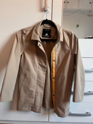 Beige trenchcoat från Didriksons - Stilren beige trenchcoat från Didriksons med rak passform och klassisk krage. Jackan har diskreta sidofickor och en gul detalj längs dragkedjan på insidan. Perfekt för dig som gillar minimalistisk och tidlös design.