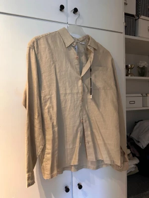 Beige linneskjorta med knappar - Stilren beige skjorta i linne med klassisk krage och knäppning framtill. Skjortan har en bröstficka och långa ärmar, perfekt för en avslappnad men snygg look. Passformen är loose och materialet känns svalt och luftigt.