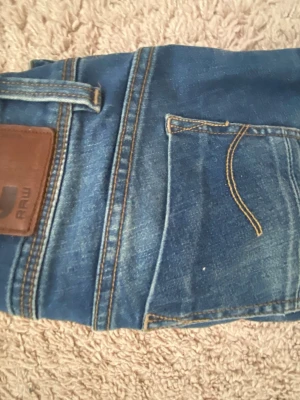 Blå jeans från G-Star RAW - Snygga blå jeans från G-Star RAW med klassisk femficksdesign och slitna detaljer. Passar nån runt 150-160 storlek 24/30 tror jag. Jeansen har raka ben och kontrastsömmar samt en brun läderpatch med logga bak i midjan. Perfekta för en avslappnad och trendig look. 