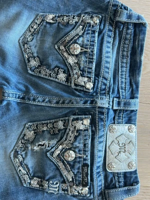 Miss Me bootcut jeans med brodyr - Snygga blå bootcut jeans från Miss Me med slitningar och broderade detaljer på bakfickorna. Jeansen har silverfärgade knappar, dekorativa sömmar och en metallplatta vid linningen. Perfekta för dig som gillar statement-jeans med unik design. Är 165 och jeansen passar mig exakt.