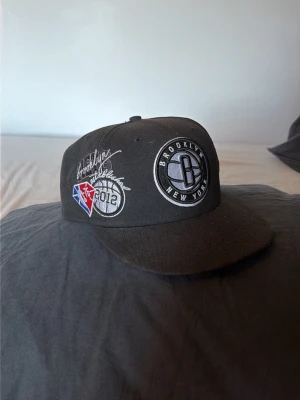 Brooklyn Nets svart keps New Era - Svart keps från New Era med Brooklyn Nets-logga framtill och broderade detaljer på sidan, inklusive NBA-logga och 2012-märke. Storlek 58,7 cm. Perfekt för dig som gillar basket och streetwear.