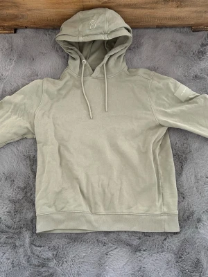 Ljusgrön hoodie Sail Racing  - Snygg ljusgrön hoodie från Sail Racing med broderad logga på huvan och patch på ärmen. Hoodien har dragsko i huvan och ribbade muddar. Tillverkad i mjuk bomull för en skön och avslappnad look.