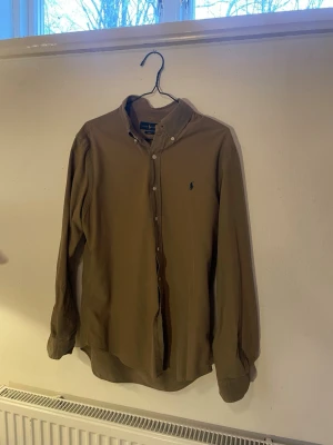 Beige skjorta från Ralph Lauren - Klassisk grön skjorta från Ralph Lauren med button-down krage och broderad logga på bröstet. Skjortan har långa ärmar och knäppning framtill. Perfekt för en stilren och avslappnad look. Hör av er vid frågor!❤️‍🩹