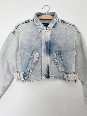 Ljusblå croppad jeansjacka från Never Denim - Snygg ljusblå croppad jeansjacka med tvättad look. Jackan har två snedställda fickor med lock och silverfärgade knappar, samt justerbara spännen i midjan. Klassisk krage och raka ärmar ger en cool och avslappnad vibe.
