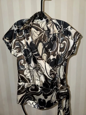 Mönstrad omlottblus med krage - Snygg omlottblus med krage och korta ärmar i glansigt material. Blusen har ett abstrakt mönster i svart, vitt och brunt med stora blommor och swirl-design. Knytband i midjan för en cool passform. Perfekt för dig som gillar statement-plagg.
