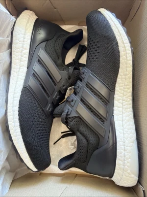 Adidas Ultra Boost svarta sneakers - Svarta Adidas Ultra Boost sneakers med stickad ovandel i mesh, klassiska tre ränder på sidan och vit Boost-sula för extra komfort. Snygg och sportig design med svart snörning och detaljer i plast. Perfekta för dig som gillar streetwear och sportig stil.
